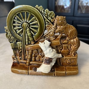 May include: A ceramic figurine featuring a spinning wheel, a dog, and a cat. The figurine is primarily brown and green with white and brown accents on the dog. The dog is reaching up towards the cat. The base has the letters "RBS".