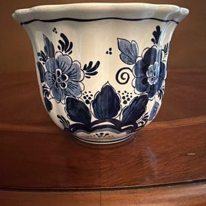 May include: A ceramic planter featuring a white base with hand-painted dark blue floral patterns and foliage. The vessel has a scalloped rim and a rounded body, decorated with intricate blue petals, leaves, and small dots arranged in a symmetrical design.
