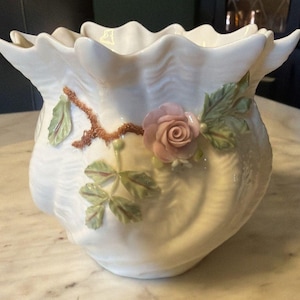 May include: A white ceramic planter with a ruffled rim and a textured surface. The planter is adorned with a delicate pink rose and green leaves, adding a touch of floral elegance. The planter is a decorative item.
