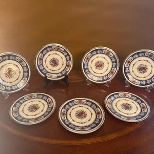 May include: A set of six round ceramic plates featuring a cream-coloured centre with a floral motif in red and blue. The wide border is decorated with a dark blue and red pattern, finished with a metallic silver rim around the outer edge.