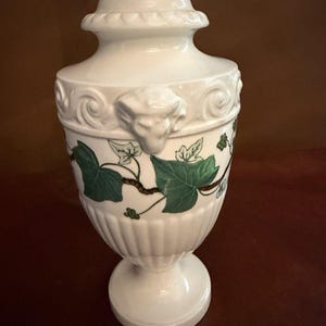 May include: A white ceramic lamp base with a fluted lower section and a smooth upper body featuring embossed ram head motifs. The surface is decorated with painted green ivy leaves and brown vines. A metallic fitting is visible at the top.