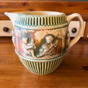 May include: A ceramic pitcher featuring a cream-colored base with green and orange accents. The exterior displays a relief pattern of figures playing musical instruments amidst trees, bordered by vertical ribbed bands at the top and bottom.