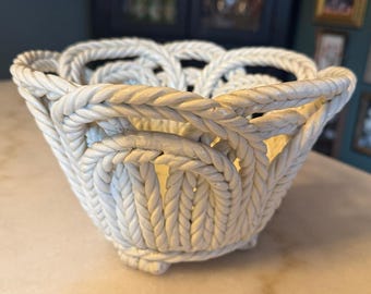 May include: A cream-colored ceramic bowl featuring a decorative openwork design that mimics the appearance of braided rope. The bowl has a rounded shape with a scalloped rim and a textured surface that replicates the twisted strands of thick cord.