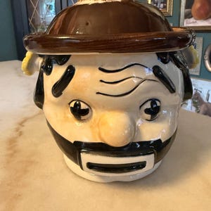 Puede incluir: Un tarro de galletas de cerámica con forma de persona que lleva un sombrero marrón. La figura tiene una cara de color crema, cejas negras, ojos negros, bigote negro y boca negra. El sombrero sirve de tapa y el tarro tiene detalles amarillos.
