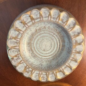 May include: A round, decorative bowl with a light blue interior and a gold-colored rim. The bowl has a scalloped edge and a concentric circle design in the center. The exterior has a distressed, antique appearance.