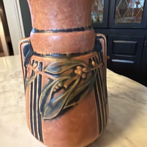May include: A terracotta-colored vase with a flared top and a cylindrical body. The vase features dark blue vertical stripes and a decorative relief of leaves and berries in shades of blue and green. The vase is a decorative home accent.