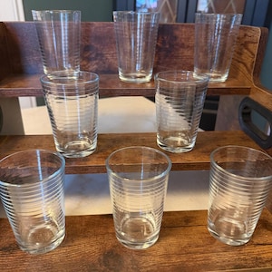 May include: A set of eight clear glass tumblers arranged on a tiered wooden display stand. Each glass features a series of thin, horizontal etched lines encircling the exterior, creating a subtle ribbed texture on the otherwise smooth surface.