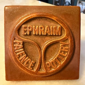 May include: A square, brown ceramic tile featuring a raised, circular emblem in the center. The text EPHRAIM is embossed at the top, with FAIENCE on the lower left and POTTERY on the lower right, all within a segmented, circular border design.