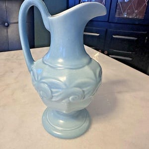 May include: A light blue ceramic pitcher with a handle and a spout. The pitcher has a decorative floral pattern around the middle and sits on a pedestal base. The pitcher is approximately 10 inches tall.
