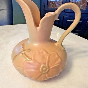 May include: Peach-colored ceramic pitcher with a bulbous base and a curved handle. The pitcher features a floral design with embossed details. The spout is wide and flared. The pitcher is likely vintage or antique.