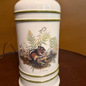 May include: A cylindrical ceramic table lamp with a glossy off-white finish and olive green horizontal bands. The body features a detailed illustration of a chipmunk surrounded by green ferns and foliage, with a signature printed at the bottom.