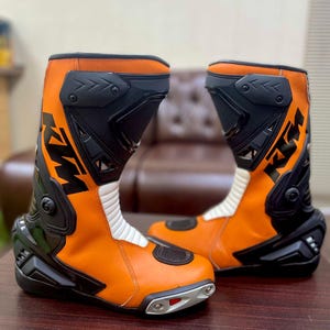 Puede incluir: Un par de botas de moto naranja y negras con el logotipo de KTM en el lateral. Las botas tienen una franja blanca en el lateral y una puntera negra.