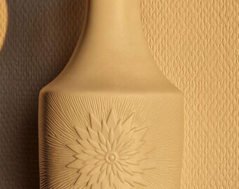 May include: A cream-coloured ceramic vase featuring a floral design. The vase has a wide base, a cylindrical body with a raised flower pattern, and a narrow neck. The flower design is centred on the body of the vase.