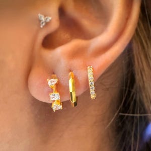 May include: Close-up of three gold hoop earrings and a small stud earring. The hoop earrings feature different designs: one with clear rectangular and oval stones, one with a geometric design, and one with small clear stones. The stud earring has a triangular design.