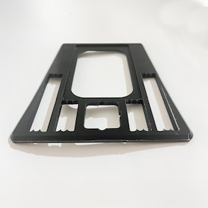 Pode incluir: Um objeto de plástico preto com uma abertura central retangular e formato trapezoidal. O objeto possui vários recortes retangulares nas laterais e na parte inferior. Colocado sobre uma superfície branca.