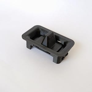 May include: A black, rectangular, 3D-printed object with a textured surface. It features a central divider and is supported by four legs. The object appears to be a car part or component.