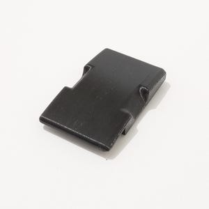 May include: A black plastic latch with a rectangular shape and a slightly curved design. The latch has two small, raised tabs on the top and bottom.