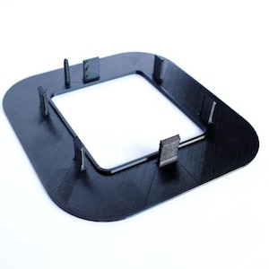 May include: A black plastic frame with a square opening and eight small tabs around the perimeter. The frame is designed to hold a square object in place.