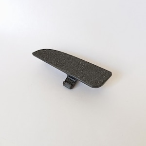 May include: A black, textured plastic part with a rectangular shape and a small, attached tab. The part has a slightly curved top surface and appears to be a component of a larger item. The background is plain white.