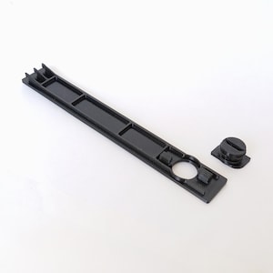May include: Black plastic component with a rectangular base, several recessed sections, and a circular opening. A separate, round, black plastic piece with a screw-like top is also present. The items are likely parts of a larger mechanism.