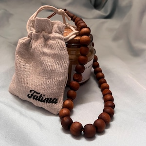 May include: A wooden bead necklace with a matching drawstring pouch. The pouch is beige and has the name "Fatima" printed on it. A small glass jar with a wooden lid is also visible.
