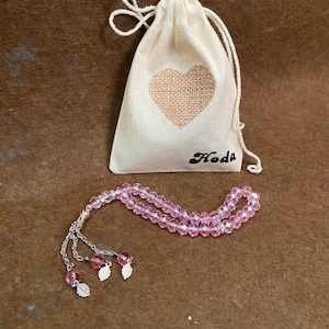 May include: Pink crystal prayer beads with a silver tassel and leaf charms, packaged in a small white drawstring bag with a heart design and the word "Hoda" printed on it.