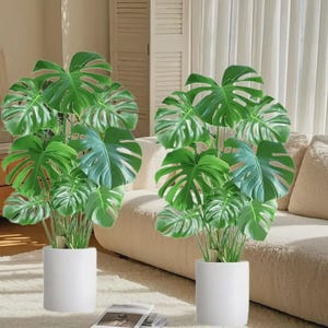 May include: Two artificial Monstera plants with large, green, split leaves in white cylindrical pots. The plants are positioned near a beige sofa and a white rug. The image is well-lit, highlighting the vibrant green of the leaves.