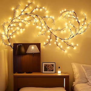 May include: A bedroom scene featuring a warm, inviting ambiance. Decorative string lights shaped like branches with small, warm white lights adorn the wall. A wooden nightstand holds lamps, a framed picture, and a small bottle.