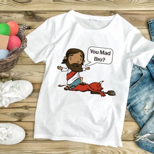Könnte beinhalten: Weißes T-Shirt mit einer Cartoon-Illustration einer Figur in einem Gewand, die auf einem roten Teufel steht. Eine Sprechblase sagt "You Mad Bro?" Das Shirt liegt auf einer Holzoberfläche mit einem Korb voller bunter Eier.