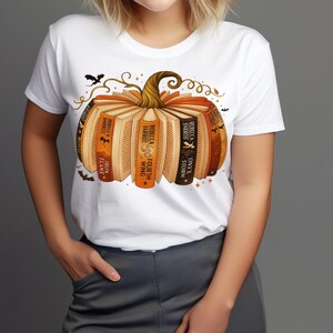 May include: White t-shirt featuring a pumpkin design made of books. The books have titles like "Iron Flame", "Fourth Wing", and "Onyx Storm". The pumpkin has a brown stem and is decorated with bats and swirls.