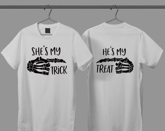 May include: Two white t-shirts hanging on a metal rod. One shirt says "She's my Trick" with a black skeleton hand graphic. The other shirt says "He's my Treat" with a matching skeleton hand graphic. The shirts are for Halloween.