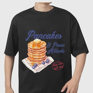 May include: Black t-shirt with a pancake design, including butter, syrup, and blueberries, with the text "Pancakes & Panic Attacks." A "buy one get one" graphic is also present. This is a casual wear item.