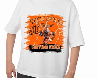 May include: White t-shirt featuring a football player in an orange uniform, with the number 19. The design includes the words "TEAM NAME" and "CUSTOM NAME" in a bold font, with lightning bolt accents and an orange and black background.