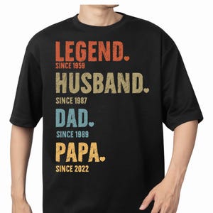 May include: Black t-shirt with the words "LEGEND," "HUSBAND," "DAD," and "PAPA" in various colors. Each word is followed by a date: 1959, 1987, 1989, and 2022, respectively. The shirt has short sleeves and a crew neck.