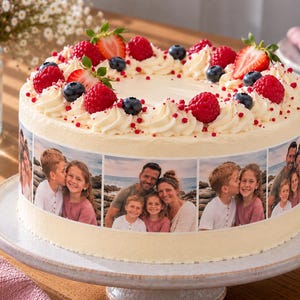 Könnte beinhalten: Ein runder Kuchen mit weißer Glasur, belegt mit frischen Erdbeeren, Himbeeren und Blaubeeren. Die Seite des Kuchens zeigt einen horizontalen Streifen mit essbaren Fotodrucken, die eine vierköpfige Familie am Strand darstellen.