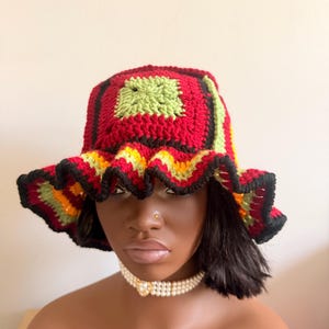 May include: A handmade crochet bucket hat with a square top and ruffled brim. The hat features a color block design with red, green, yellow, and black yarn. The hat is a stylish accessory.