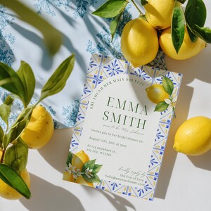 May include: A bridal shower invitation featuring a lemon theme. The invitation has a white background with blue and yellow decorative borders. The text reads "Emma Smith" and includes event details. Fresh lemons and leaves are arranged around the invitation.