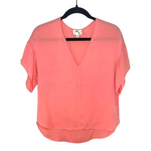 May include: A coral-colored short-sleeve blouse with a V-neck. The blouse is hanging on a black hanger. The brand name and size 4 are visible on the label. The fabric appears to be lightweight and flowing.