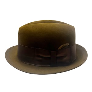 May include: A brown fedora hat with a dark brown band and bow. The hat features a classic design with a slightly curved brim and a pinched crown. A small feather is tucked into the band.