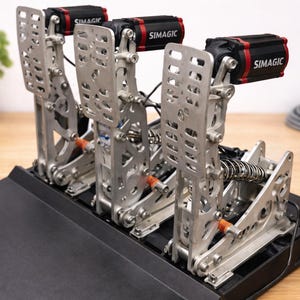 May include: A silver racing simulator pedal set with three pedals. Each pedal has a SIMAGIC motor with a black and red casing. The pedals are mounted on a black base. The background includes a plant in a white pot and a coiled cable.