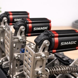 Op de afbeelding: Close-up van een sim racing pedaalset. De pedalen zijn gemaakt van zilverkleurig metaal met zwarte en rode accenten. De tekst "SIMAGIC" is zichtbaar op de zwarte cilinders. De pedalen zijn gemonteerd op een zwarte basis.