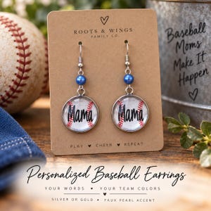 May include: A pair of silver-toned dangle earrings on a tan display card. Each earring features a round charm with a baseball pattern and the word Mama in black script, topped with a single blue bead. The card displays the text Roots & Wings Family Co. and Play Cheer Repeat.