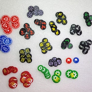 May include: A collection of colorful game tokens in various shapes and designs. The tokens feature symbols like arrows, lightning bolts, and numbers, in red, blue, green, yellow, black, and white. These tokens are likely used for board games or tabletop games.