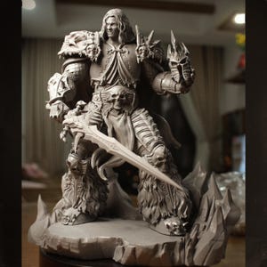 May include: A detailed grey statue of a warrior figure, holding a large sword. The statue features intricate armour with skull details, long flowing hair, and a cape. The figure stands on a rocky base, showcasing a fantasy theme.