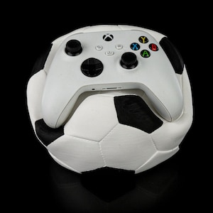 May include: A white Xbox controller sits on a football. The controller has black joysticks and coloured buttons. The football is white with black pentagons.