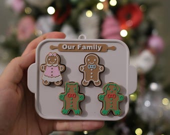 May include: A gray, rectangular ornament with the words "Our Family" and a rolling pin design. It features four gingerbread people: a girl in a pink dress, a man with a bow tie, and two gingerbread pets. The ornament is held in front of a Christmas tree.
