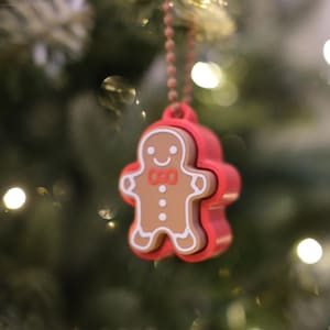 Op de afbeelding: Een kerstboomversiering met een peperkoekmannetje. De bruine ornament met witte details en een rode strik is ingekapseld in een rode lijst. Het hangt aan een kralenketting tegen een vage kerstboomachtergrond.