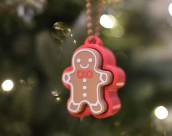 May include: A festive gingerbread man Christmas ornament. The ornament is brown with white accents and a red bow tie, encased in a red frame. It hangs from a beaded chain against a blurred Christmas tree background.