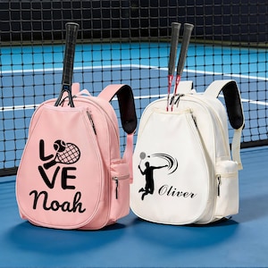 Könnte beinhalten: Zwei Tennisrucksäcke, einer rosa und einer weiß, werden vor einem blauen Platzhintergrund gezeigt. Der rosa Rucksack hat die Worte "LOVE Noah" und eine Tennisgrafik. Der weiße Rucksack zeigt eine Badmintonspieler-Grafik und den Namen "Oliver". Beide haben schwarze Träger.
