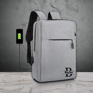 May include: A gray backpack with a black handle and straps. The backpack has a front pocket with a zipper and the name "Baylor" printed on the front. A smartphone is charging next to the backpack.